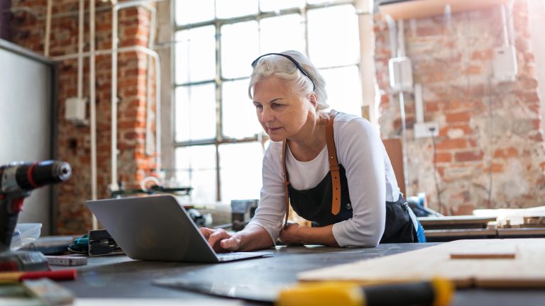 Frau mit grauen Haaren sitzt in Werkstatt vor dem Laptop
