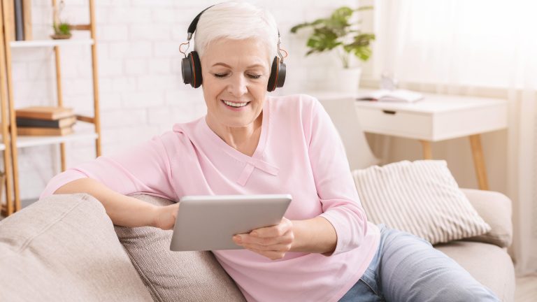 Frau mit grauen Haaren sitzt auf Sofa. Sie trägt große Kopfhörer und benutzt ein Tablet.